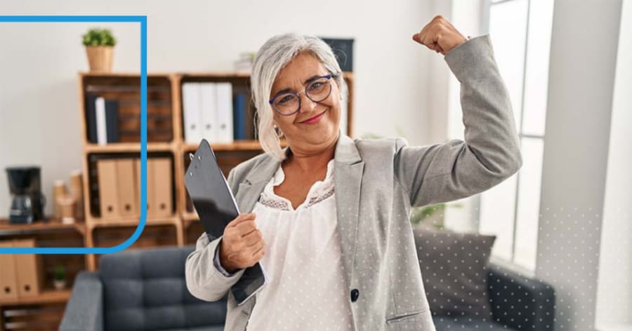 A woman with grey hair wearing a grey blazer is holding her arm up, showing strength.